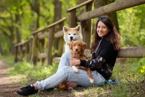 Kobieta z dwoma psami w lesie. Jeden piesek jest na jej kolanach, drugi za nim. Wskazówki, jak uspokoić sukę podczas cieczki, mogą być pomocne.