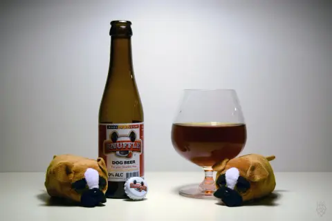 Dwa pluszowe pieski patrzą na butelkę piwa "Snuffle Dog Beer" i kieliszek. Czy pies może pić piwo? To piwo jest dla psów, 0% alkoholu.